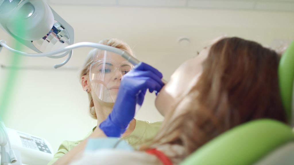 Female dentist treating patient teeth. Blonde doctor in protective goggles